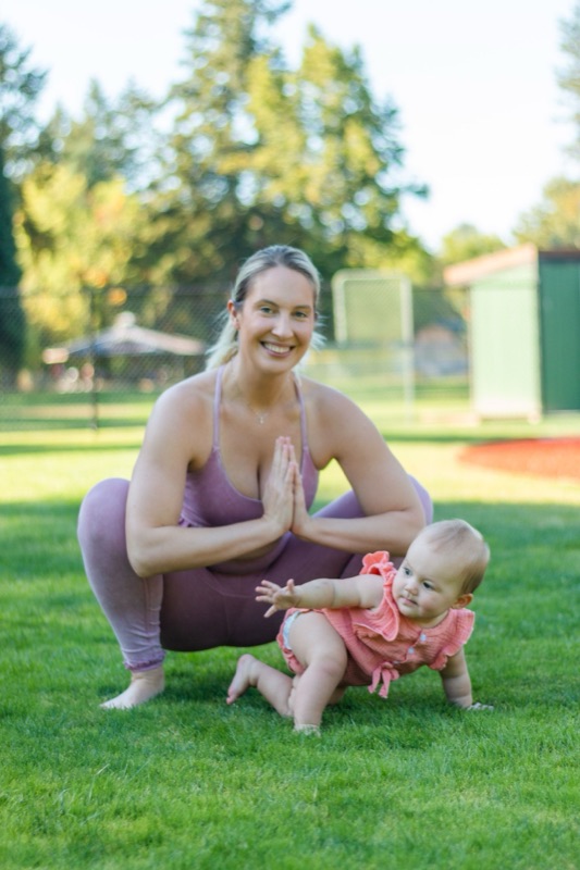 A mother in a yoga squat pose smiling at her crawling baby on the grass